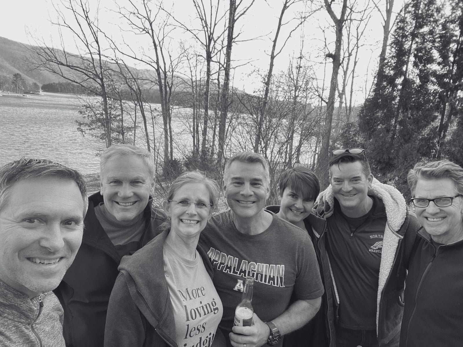 The seven members of James Doak Connection smiling in a group selfie by a lake, bare winter trees behind them.